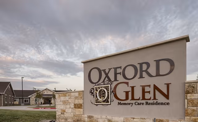 Stone entrance sign reading "Oxford Glen Memory Care Residence" with the facility building and cloudy sky in the background.