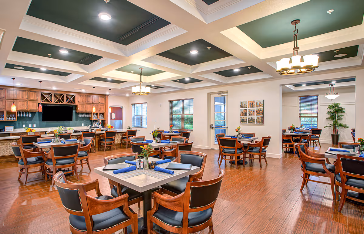 A spacious dining room with multiple wooden tables and chairs arranged neatly. Each table is set with blue napkins and small flower vases. The room features a coffered ceiling with recessed lighting and hanging light fixtures. There is a bar area with wooden cabinets, wine racks, and a mounted flat-screen TV. Large windows and a glass door allow natural light to enter the room.