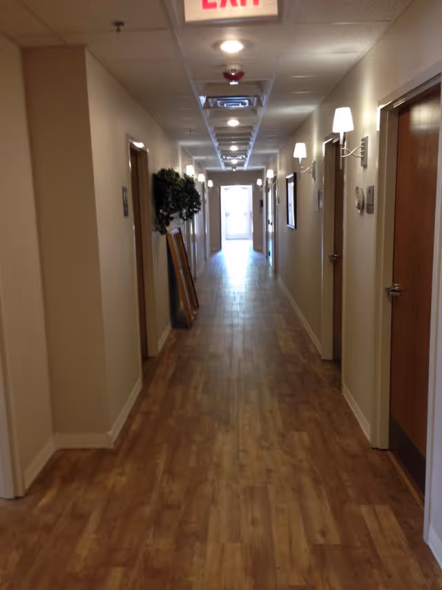 A long, well-lit hallway with wooden flooring and beige walls. Several closed wooden doors line both sides of the corridor, each with a wall-mounted light fixture above. There is an exit sign at the ceiling near the foreground and a bright doorway at the far end of the hallway. A plant and a framed item are placed against the left wall.
