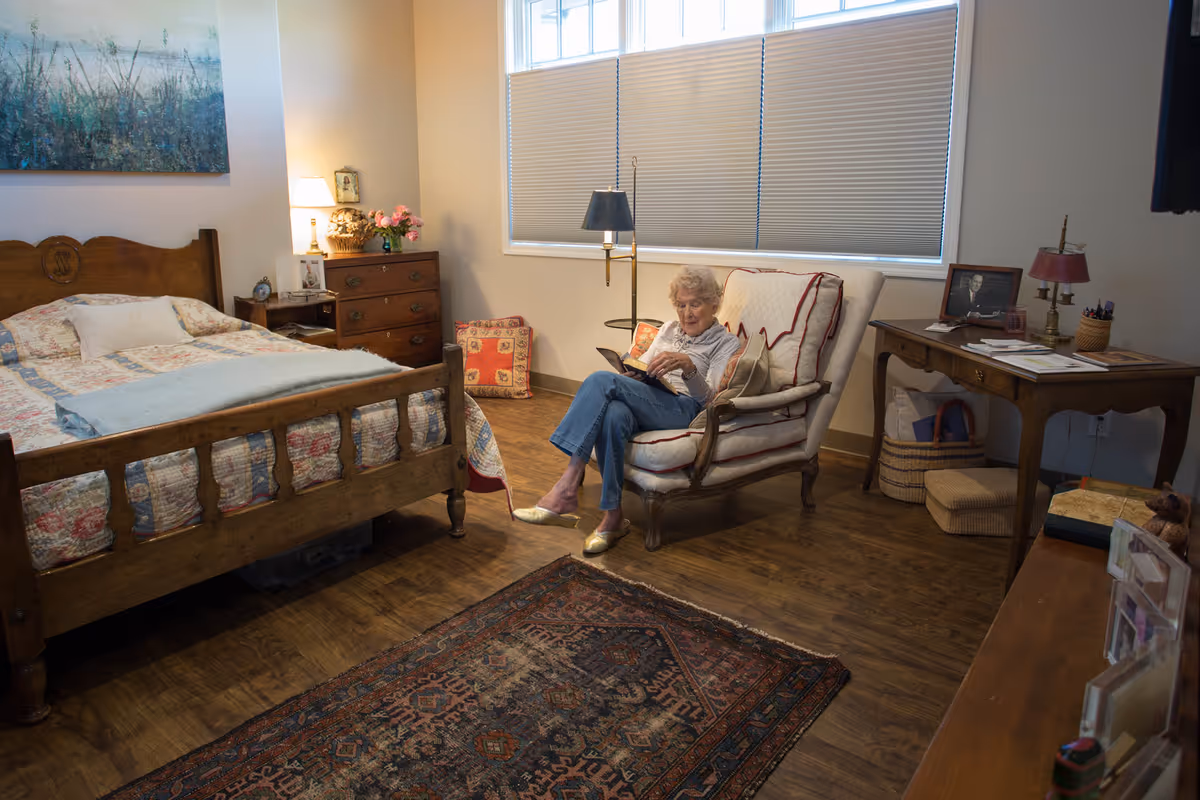 An elderly woman sits reading in a cozy bedroom with a wooden bed, dresser, armchair and rug.