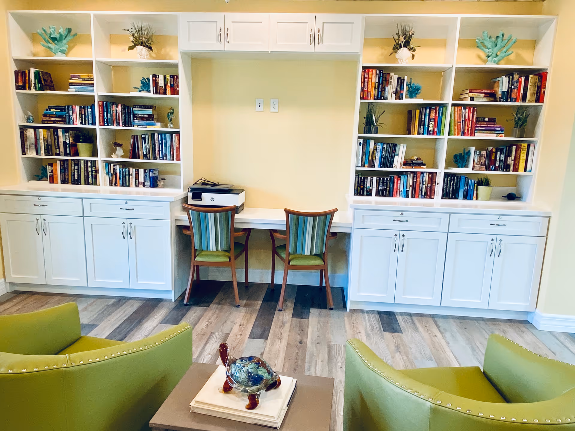 A cozy reading and work area in an assisted living facility featuring white built-in bookshelves filled with books and decorative items. Two striped chairs are placed at a built-in desk with a printer on it. In the foreground, there are two green armchairs and a small table with a decorative glass turtle on top.