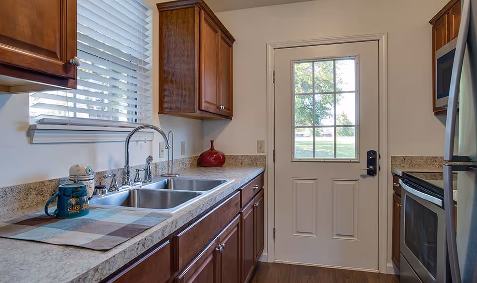 A kitchen interior featuring wooden cabinets, a double stainless steel sink, a window with white blinds above the sink, a countertop with a red vase and two mugs, and a white door with a window leading outside. Stainless steel appliances including a microwave and oven are visible on the right side.