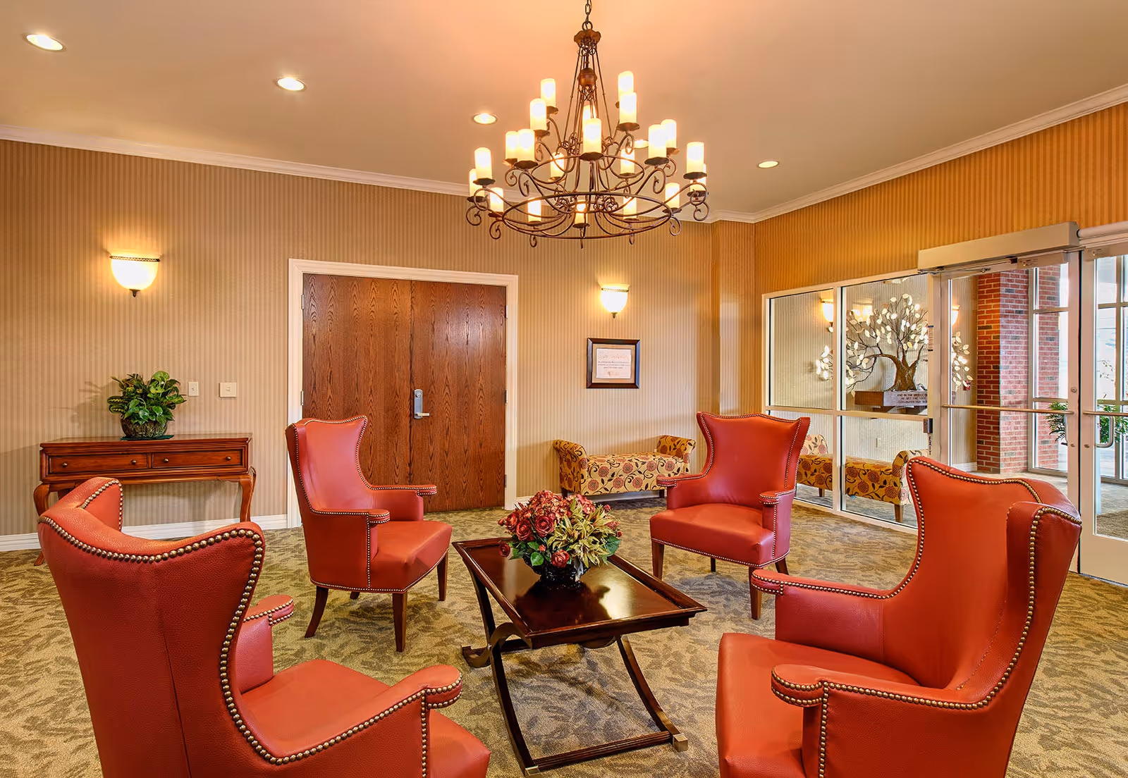 A cozy sitting area with four red leather armchairs arranged around a dark wooden coffee table with a floral centerpiece. The room has beige striped wallpaper, a green patterned carpet, a wooden console table with a plant, a chandelier with multiple lights, and a large glass door leading outside.