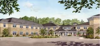 Exterior view of a two-story assisted living facility building with a large entrance canopy, surrounded by trees and greenery under a clear sky.
