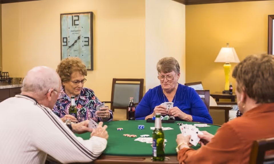 Four elderly people sitting around a green poker table playing cards and poker chips, with drinks on the table in a warmly lit room with yellow walls and a large clock on the wall.