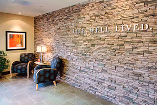 Stone accent wall with 'LIVE WELL LIVED' slogan, chairs, lamp, and framed art in a lobby.