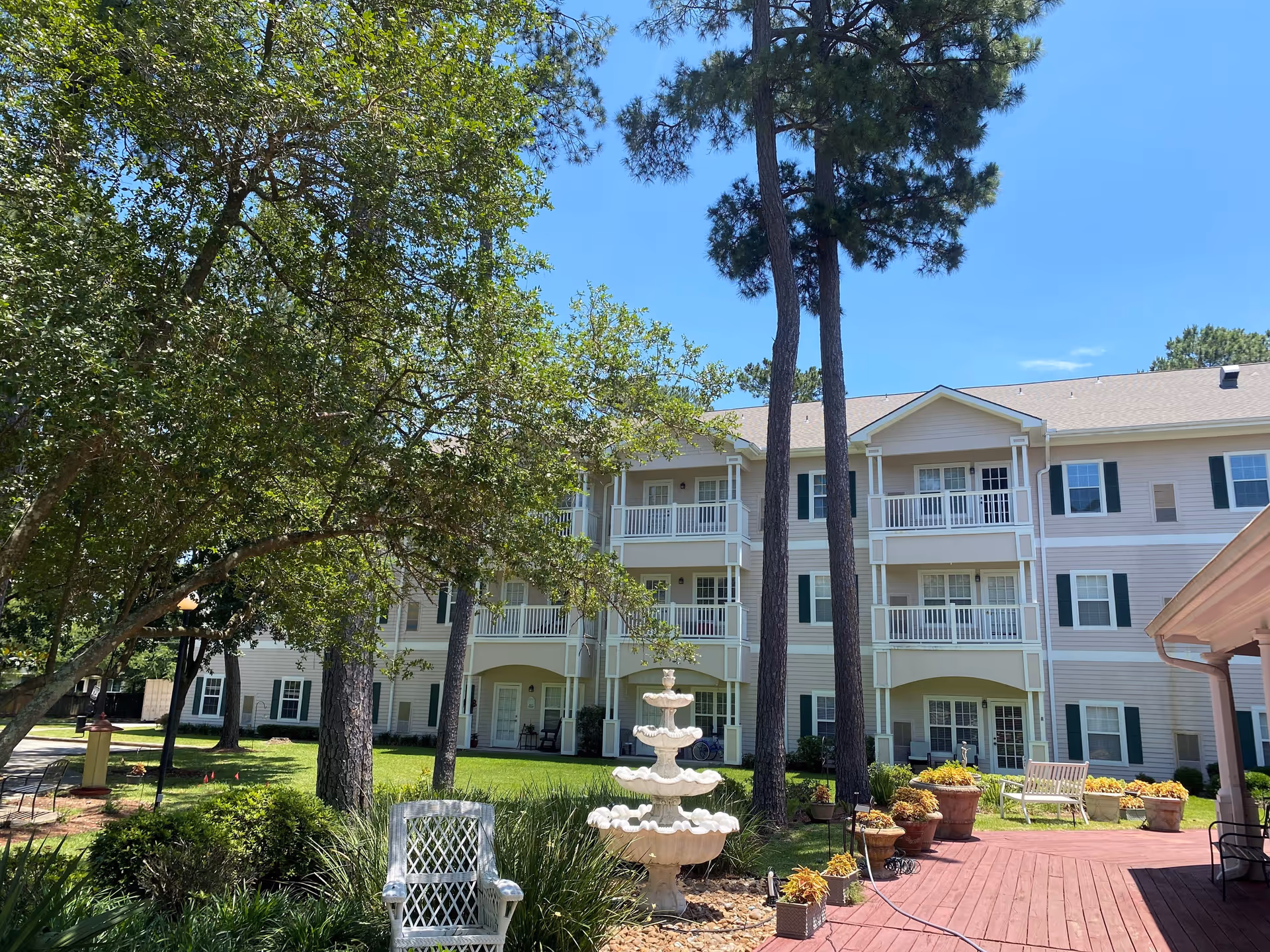 Outdoor garden area at King's Preserve at Kingwood featuring a multi-tiered white fountain, white wicker chair, potted plants, tall trees, and a three-story building with balconies and windows in the background under a clear blue sky.