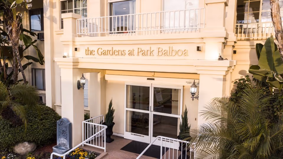 Entrance of The Gardens at Park Balboa facility with double glass doors, potted plants on either side, outdoor wall lanterns, and surrounding greenery including palm trees and bushes.