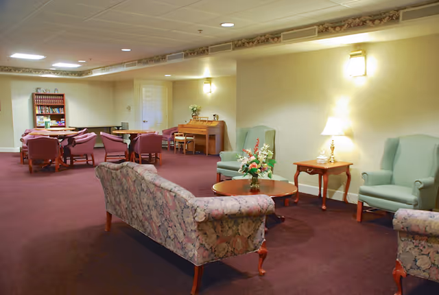 A cozy senior living facility common area with floral patterned sofas, green armchairs, wooden tables, a lamp, and a piano against the wall. The room has soft lighting and a carpeted floor.