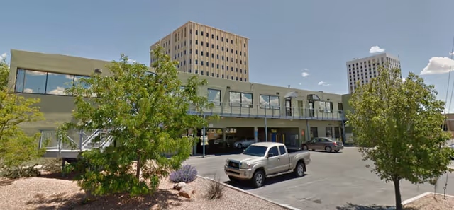 Exterior view of a two-story senior living facility building with a parking lot in front. There are several trees and shrubs in the landscaped area near the building. Two taller buildings are visible in the background under a partly cloudy sky.