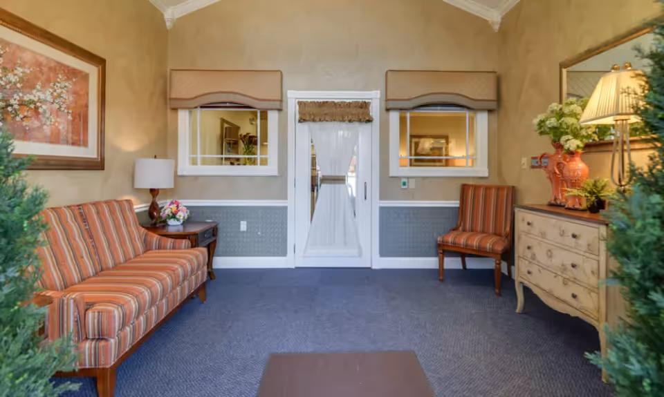 Small reception room with striped sofa and chairs, side tables and lamps, decorative plants, and a central white door with windows.