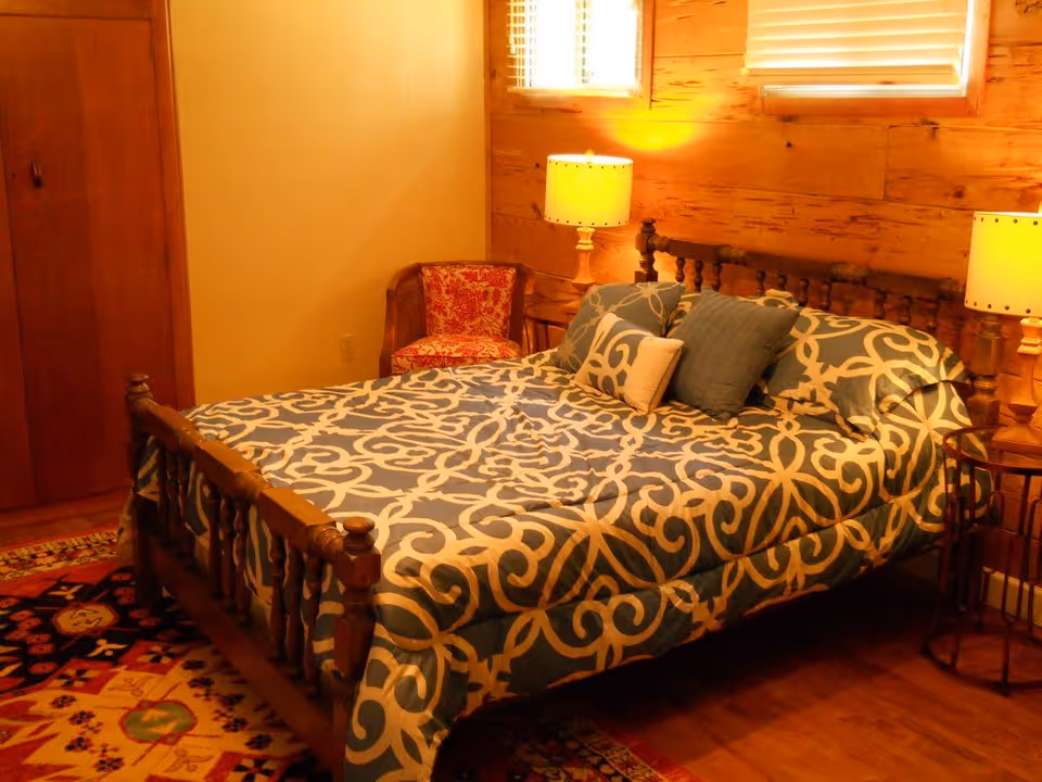 A cozy bedroom with a wooden bed frame and a blue and white patterned bedspread. The bed has several pillows, and there are two bedside tables each with a lamp. A red patterned chair is placed in the corner near a wooden closet. The room has wooden walls and a window with blinds letting in natural light.