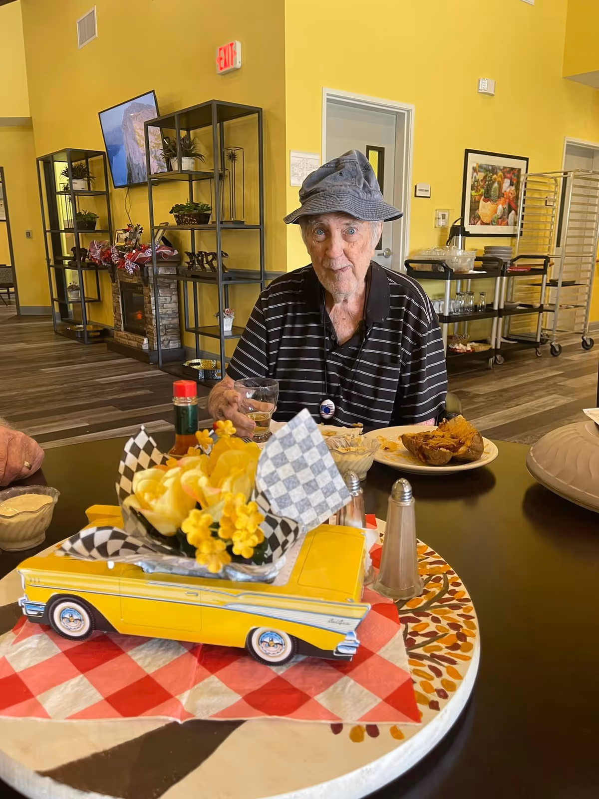 An elderly man wearing a bucket hat sits at a table with plates and a yellow toy-car floral centerpiece in a bright dining room.