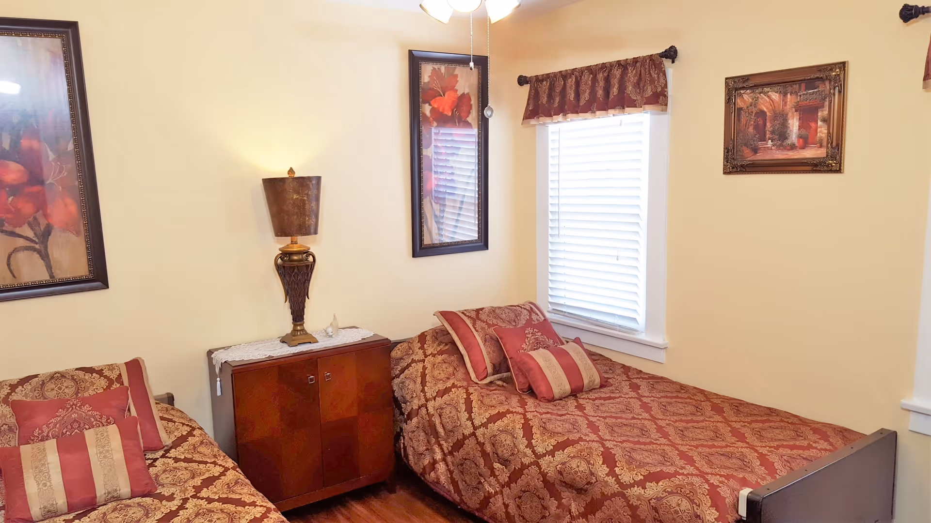 A bedroom with two twin beds covered in matching red and gold patterned bedspreads and pillows. Between the beds is a wooden nightstand with a decorative lamp and a lace runner. The walls are painted light yellow and decorated with framed artwork. A window with white blinds and a red valance is on the right wall.