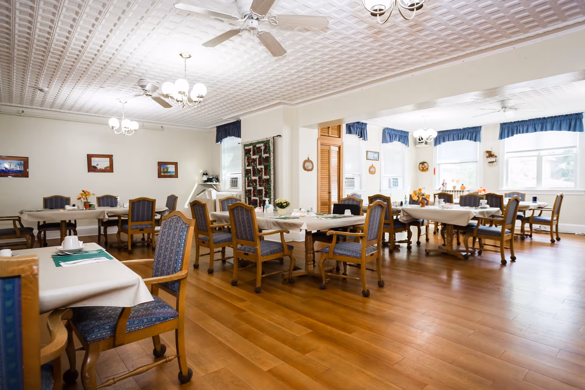 A bright dining room with wooden floors and multiple tables covered with beige tablecloths. Each table is surrounded by wooden chairs with blue patterned cushions. The room has white walls decorated with framed pictures and a quilt hanging on one wall. There are large windows with blue valances letting in natural light, ceiling fans, and chandeliers providing additional lighting.