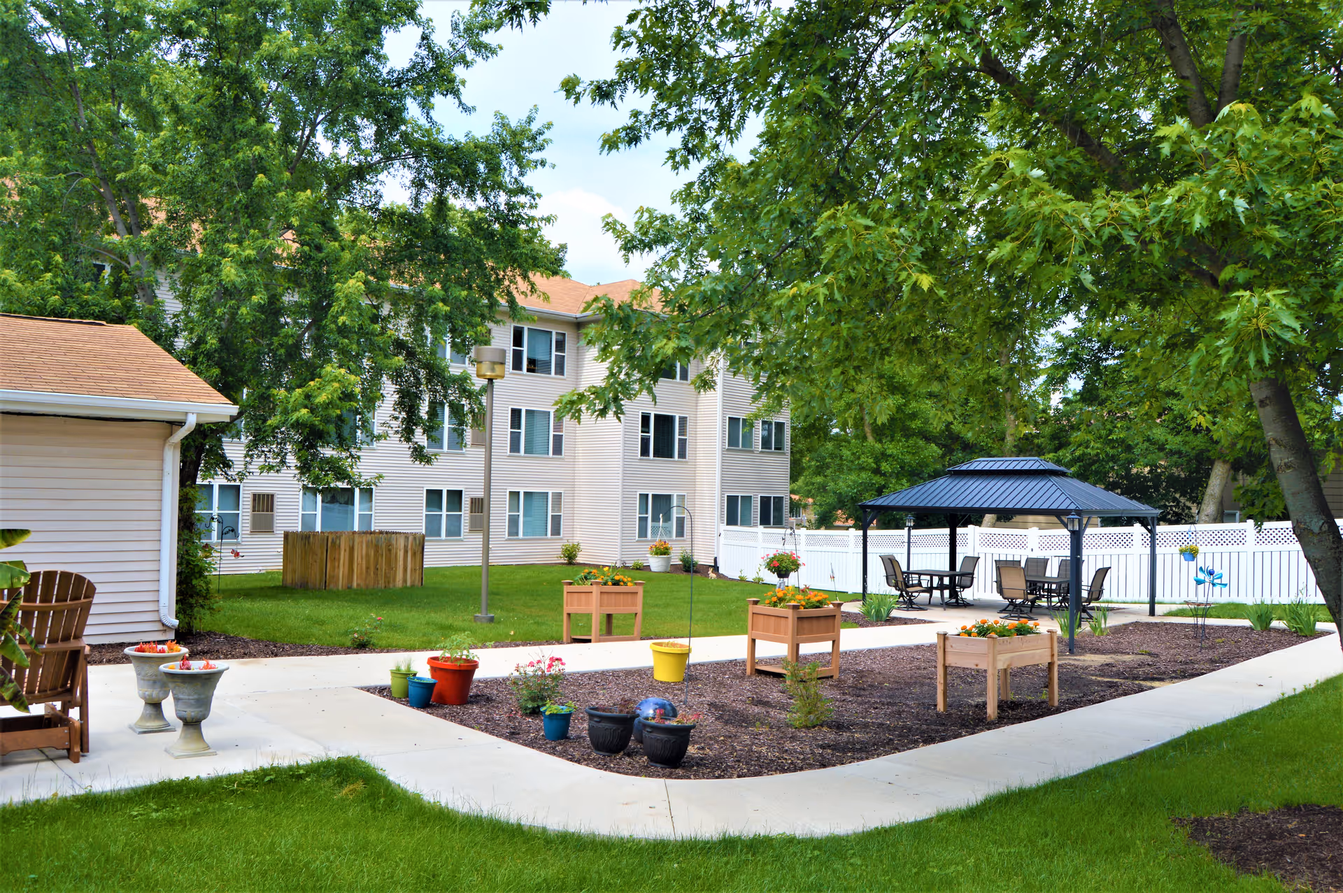 Outdoor garden area at Garnett Place featuring a paved walkway, several flower pots and raised garden beds with plants, a gazebo with tables and chairs underneath, surrounded by green grass and trees, with a multi-story residential building in the background.