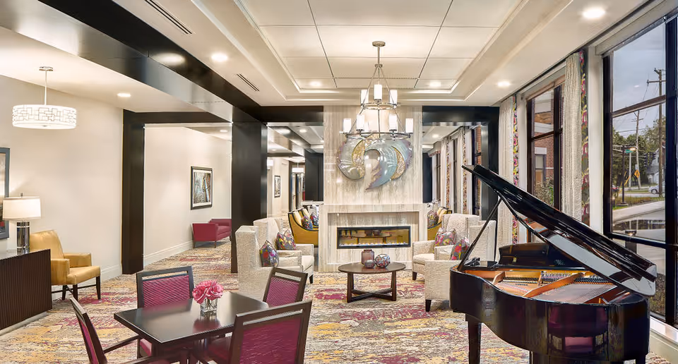 A spacious and well-lit living room area featuring a grand piano on the right side near large windows. The room has multiple seating arrangements with beige armchairs and colorful cushions around a central fireplace adorned with modern art. There are also tables with chairs, a patterned carpet, and decorative lighting fixtures on the ceiling.