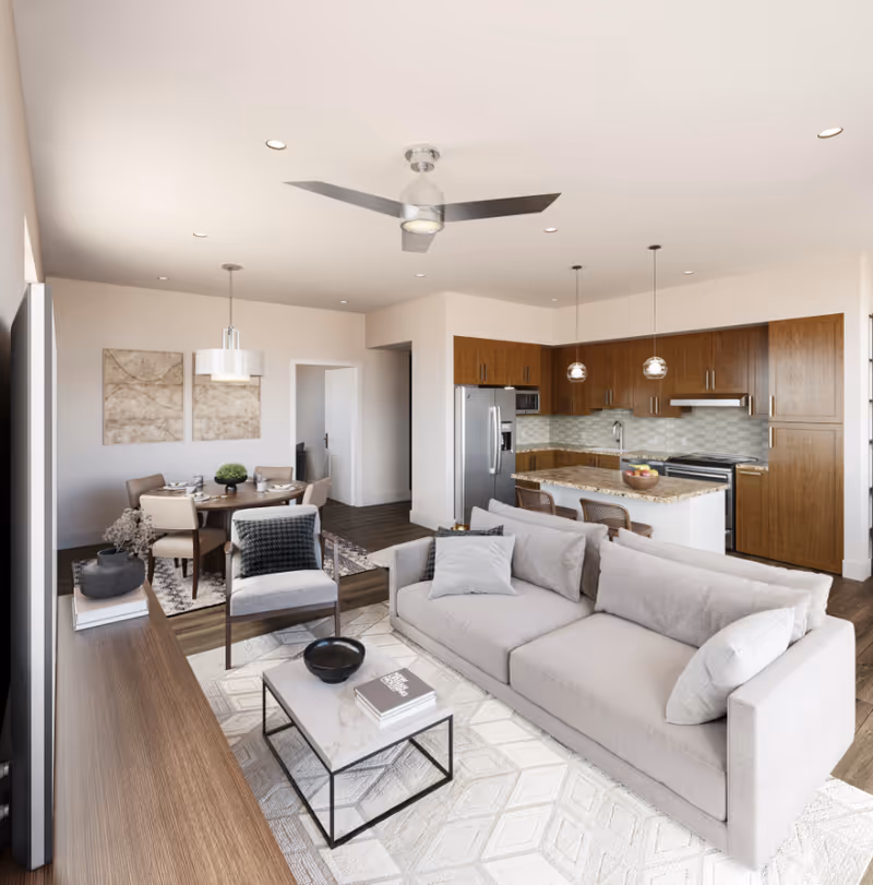 A modern open-concept living area featuring a light gray sofa with cushions, a small marble coffee table with books and a bowl, a wooden TV console, and a single armchair with a dark cushion. In the background, there is a dining area with a round wooden table and four chairs, two abstract wall art pieces, and a kitchen with wooden cabinets, stainless steel appliances, a kitchen island with a bowl of fruit, and pendant lights.