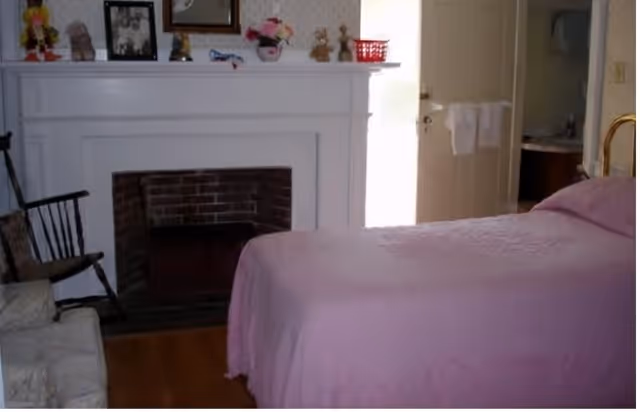 A bedroom with a bed covered in a pink bedspread, a white fireplace with decorative items on the mantel, a wooden rocking chair, and an armchair. A door is open leading to a bathroom with a sink and towels visible.