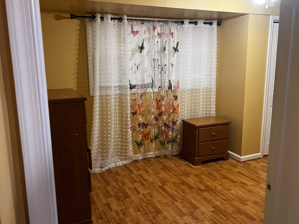 A small room with wooden flooring and yellow walls. There is a window covered with sheer white curtains decorated with colorful butterfly patterns. To the left is a wooden dresser, and to the right is a small wooden nightstand with two drawers. The room is viewed through a doorway with white trim.
