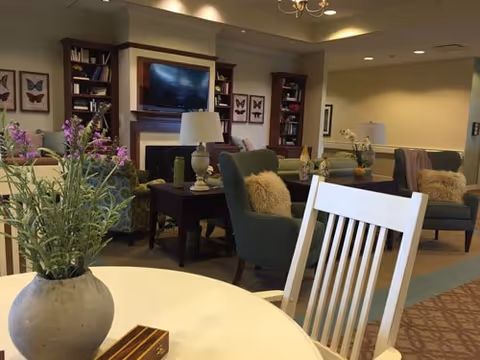 A cozy living room area in a senior living facility with comfortable green armchairs, a white wooden chair, a round table with a vase of purple flowers, bookshelves, a flat-screen TV mounted above a fireplace, and framed butterfly artwork on the walls.