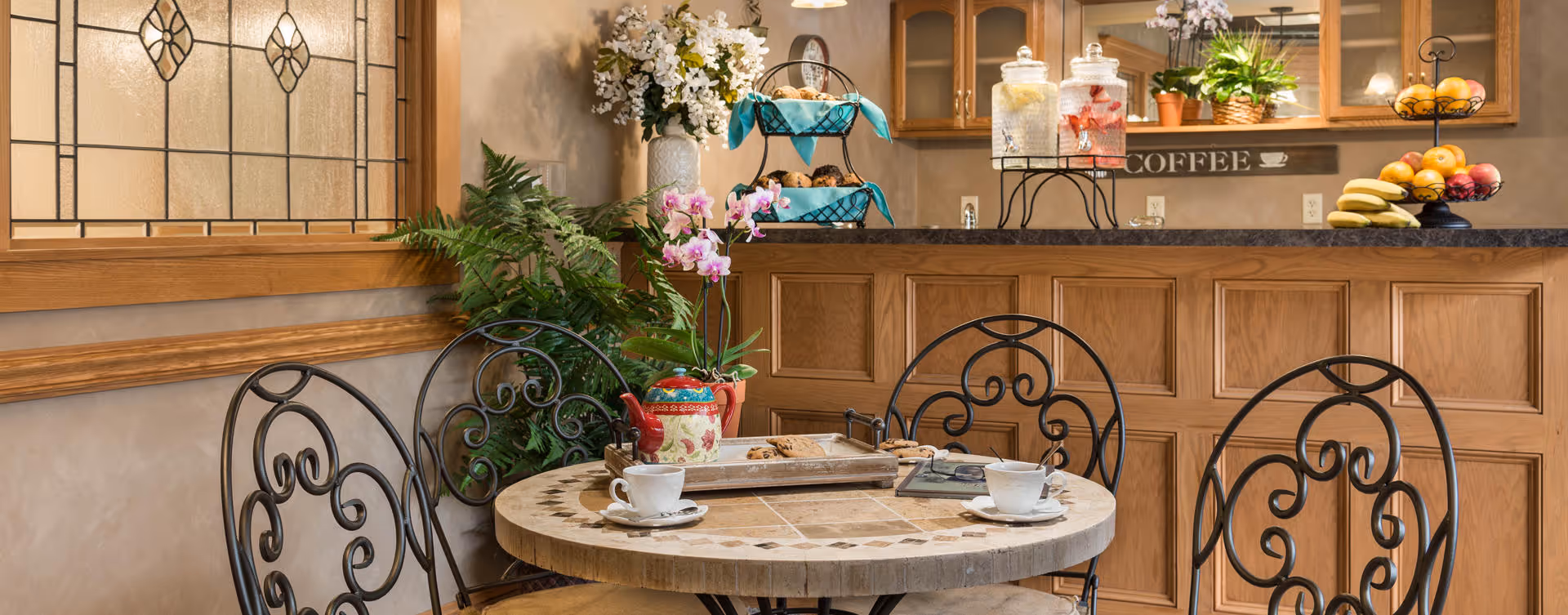 A cozy dining area with a round table and four ornate metal chairs. On the table are two white coffee cups on saucers, a tray with cookies, a decorative teapot, and a small potted orchid. Behind the table is a counter with glass dispensers containing beverages, a tiered tray with pastries, and a fruit stand with bananas, oranges, and apples. The background includes wooden cabinetry, a mirror, and a sign that reads 'COFFEE'. There are also decorative plants and a stained glass window on the left wall.