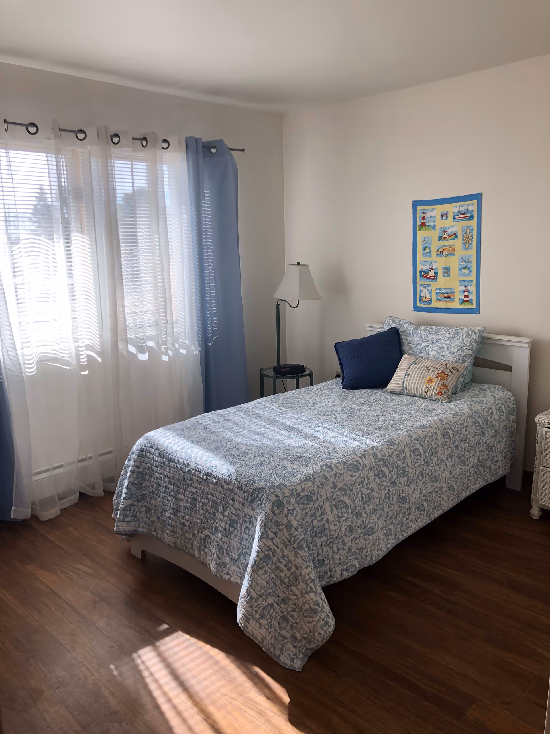 A bright bedroom with a single bed covered in a blue and white patterned quilt and three pillows. There is a window with sheer white curtains and blue drapes allowing sunlight to enter the room. A small bedside table with a lamp and a phone is next to the bed. A colorful nautical-themed wall hanging is displayed above the headboard. The floor is wooden.