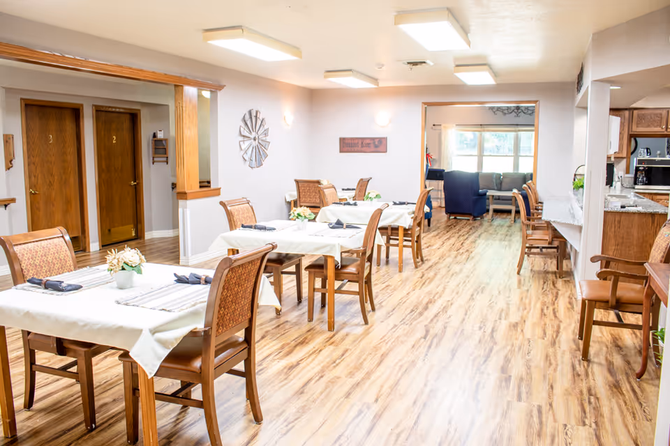 A bright dining area in a senior living facility with several tables covered in white tablecloths, each set with napkins and small flower arrangements. The room has wood flooring and light-colored walls. In the background, there is a living room area with a couch and armchairs near large windows. To the right, part of a kitchen area with wooden cabinets and a countertop is visible. Two wooden doors labeled '2' and '3' are on the left side of the image.