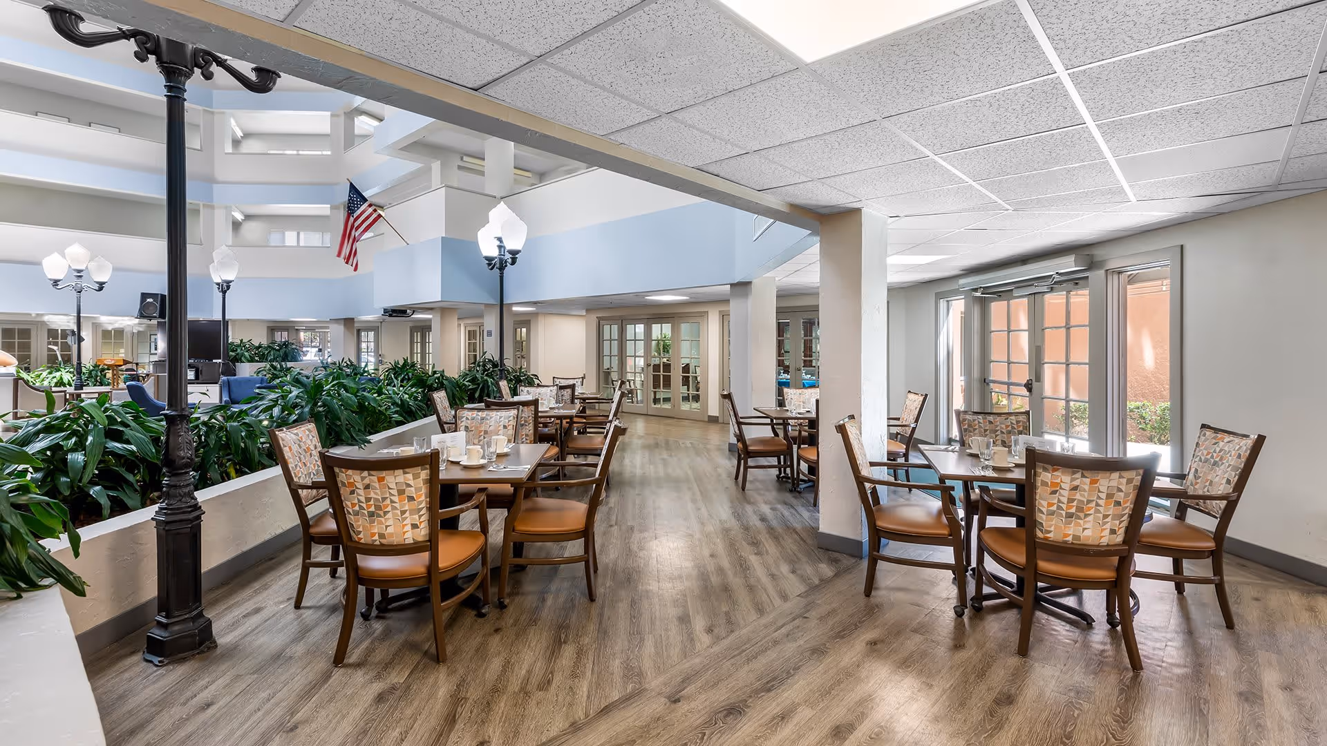 A spacious dining area in a senior living facility with multiple wooden tables and chairs arranged neatly. The room features wood flooring, large windows letting in natural light, indoor plants along a divider, and decorative streetlamp-style lights. An American flag is visible in the background near a multi-story atrium.
