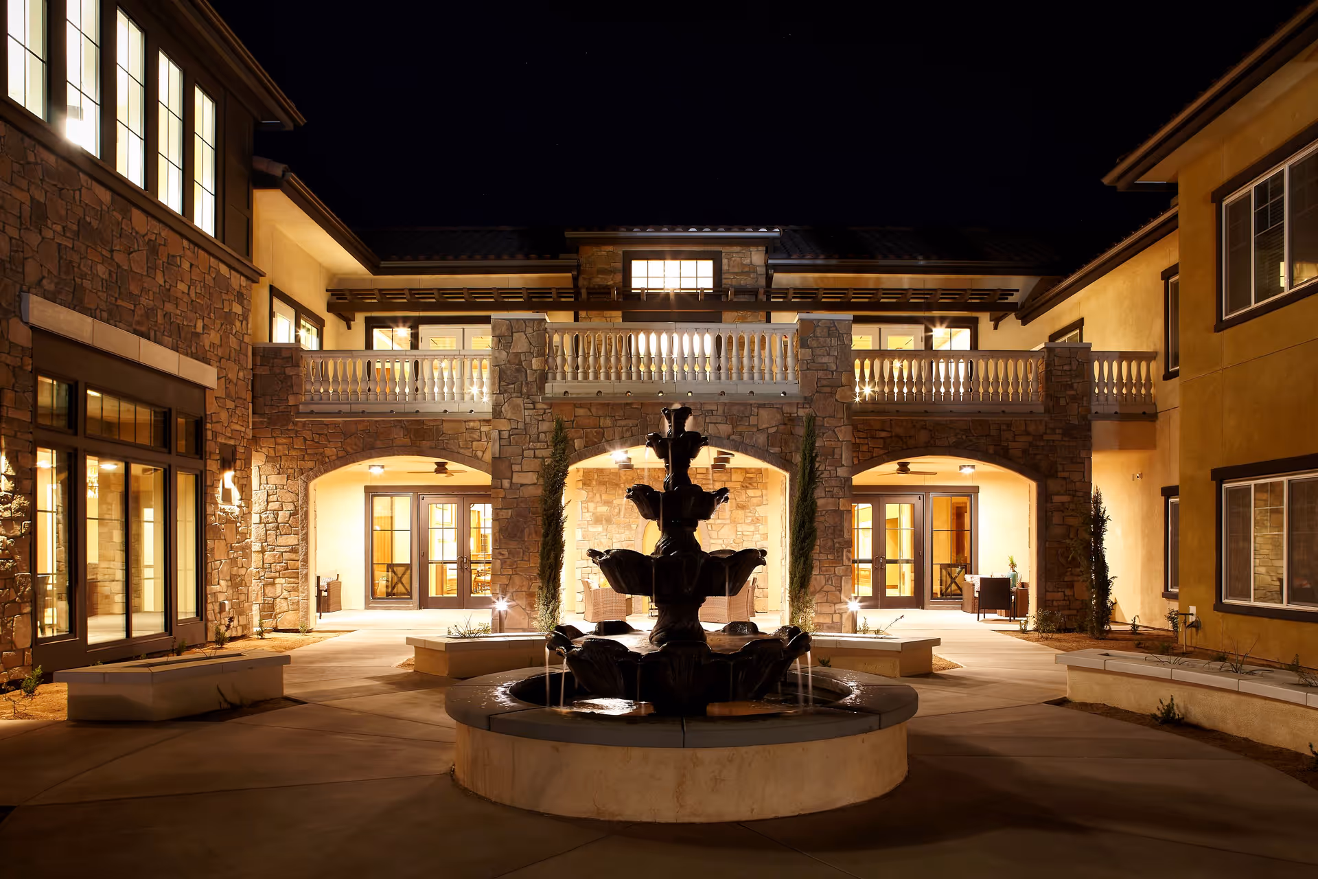Night view of an elegant courtyard at Bella Villaggio facility featuring a multi-tiered water fountain in the center, surrounded by stone and stucco buildings with illuminated windows and arched doorways, and a balcony with white balustrades above.