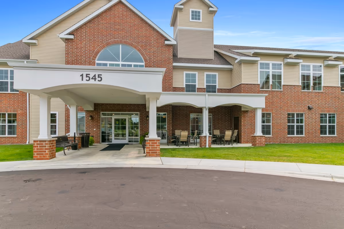 Front exterior view of a senior living facility named Gracepointe Crossing Gables, featuring a brick and beige siding building with large windows, a covered entrance with the number 1545 above it, benches, and outdoor seating areas on a concrete patio.
