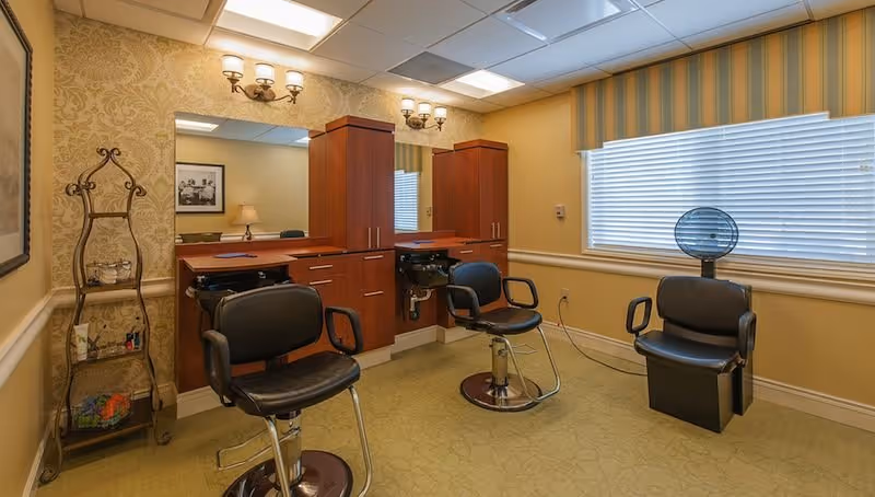 Small salon-style interior with three styling chairs, wooden cabinets and mirrors, and a hooded hair dryer by a window.