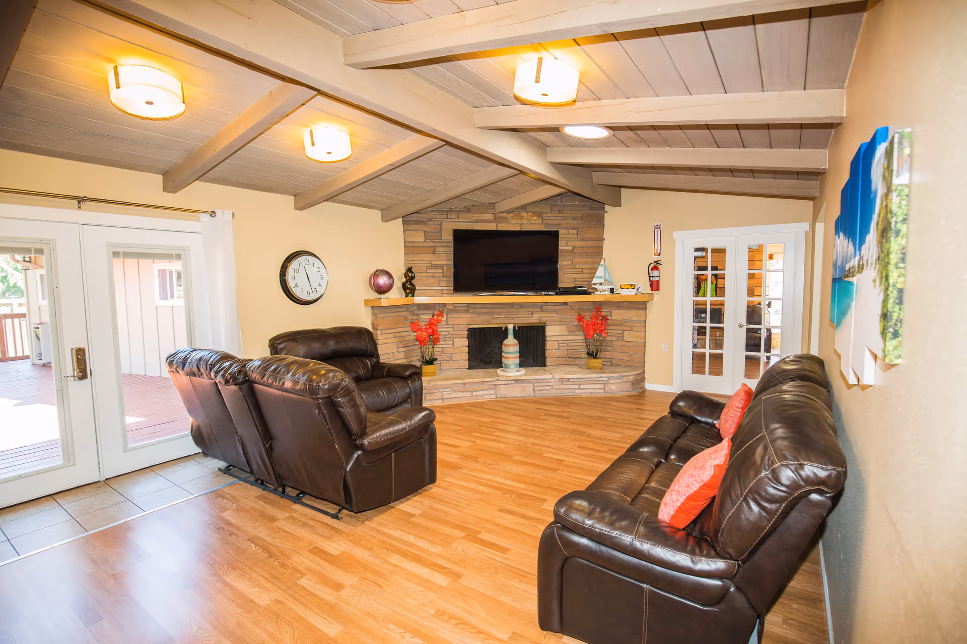 A cozy living room with wooden flooring, beige walls, and a vaulted ceiling with exposed beams. The room features two dark brown leather sofas with red cushions, a stone fireplace with decorative items on the mantel, and a flat-screen TV mounted above it. There is a clock on the wall, a fire extinguisher near a set of French doors, and large glass doors leading to an outdoor deck.