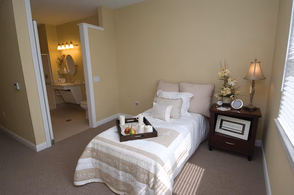 A small bedroom with a single bed covered in a beige and white quilt, adorned with multiple pillows. A wooden nightstand beside the bed holds a lamp, a clock, a framed picture, and a vase with flowers. A tray with candles and fruit is placed on the bed. The room has beige walls and carpeted floor. An open doorway leads to a bathroom with a sink, mirror, and toilet visible.