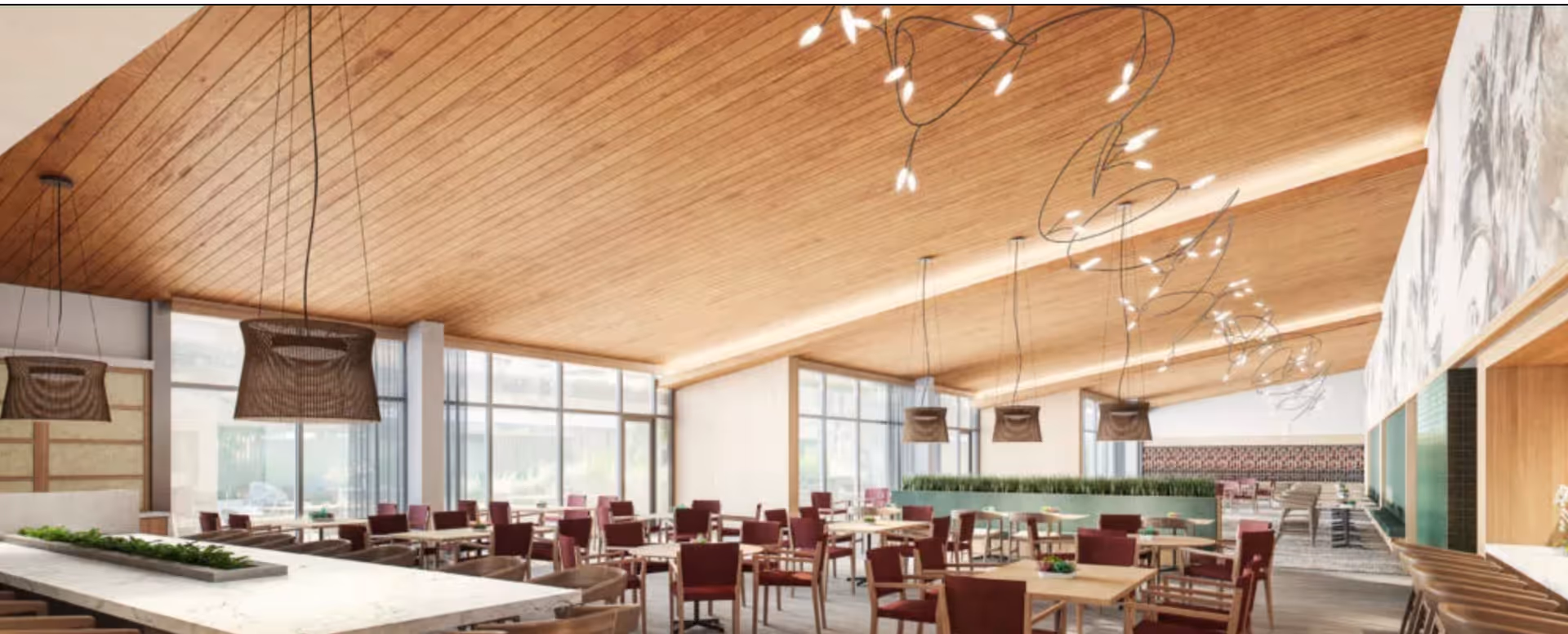 A spacious dining room with wooden ceiling and floor-to-ceiling windows allowing natural light. The room features multiple wooden tables with red cushioned chairs arranged neatly. Modern pendant lights hang from the ceiling, and there are green plants placed on a central divider and a marble countertop on the left side.