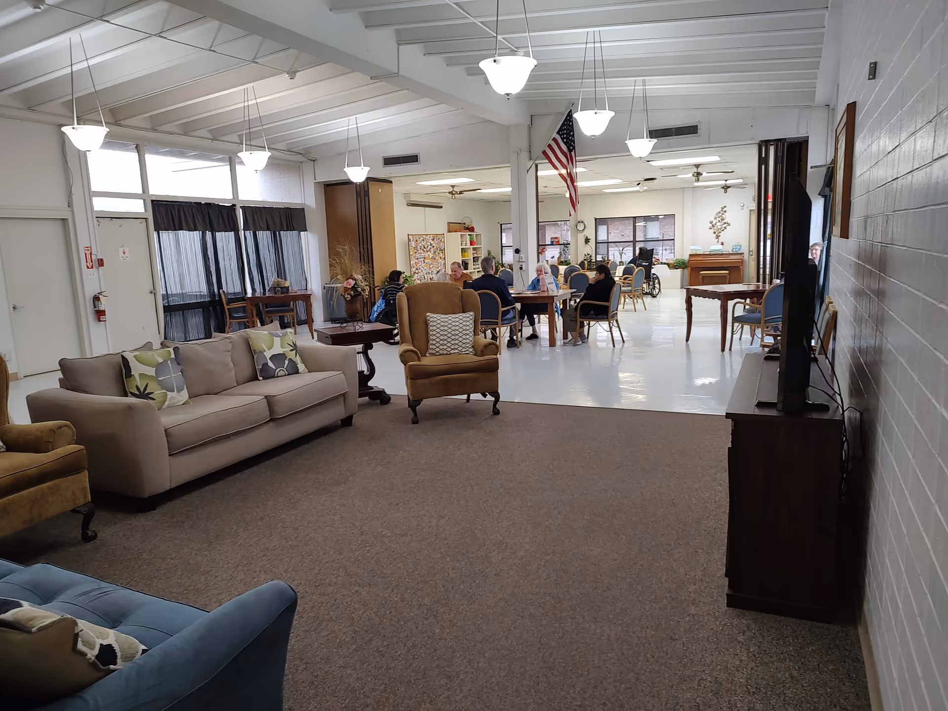 Open communal living area with sofas and armchairs in the foreground and tables with seated residents in the background.