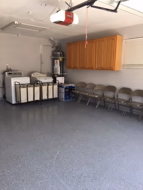A laundry room with two washing machines and a laundry sorter with multiple bags. There are wooden cabinets mounted on the wall above a row of folding chairs. A water heater is visible in the corner, and a garage door opener is mounted on the ceiling.