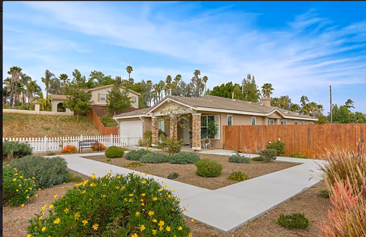 Single-story residential building with a front porch supported by stone pillars, surrounded by a landscaped garden with bushes and yellow flowers. A white picket fence and a wooden fence enclose the area, with a concrete walkway leading to the entrance. Palm trees and other houses are visible in the background under a partly cloudy blue sky.