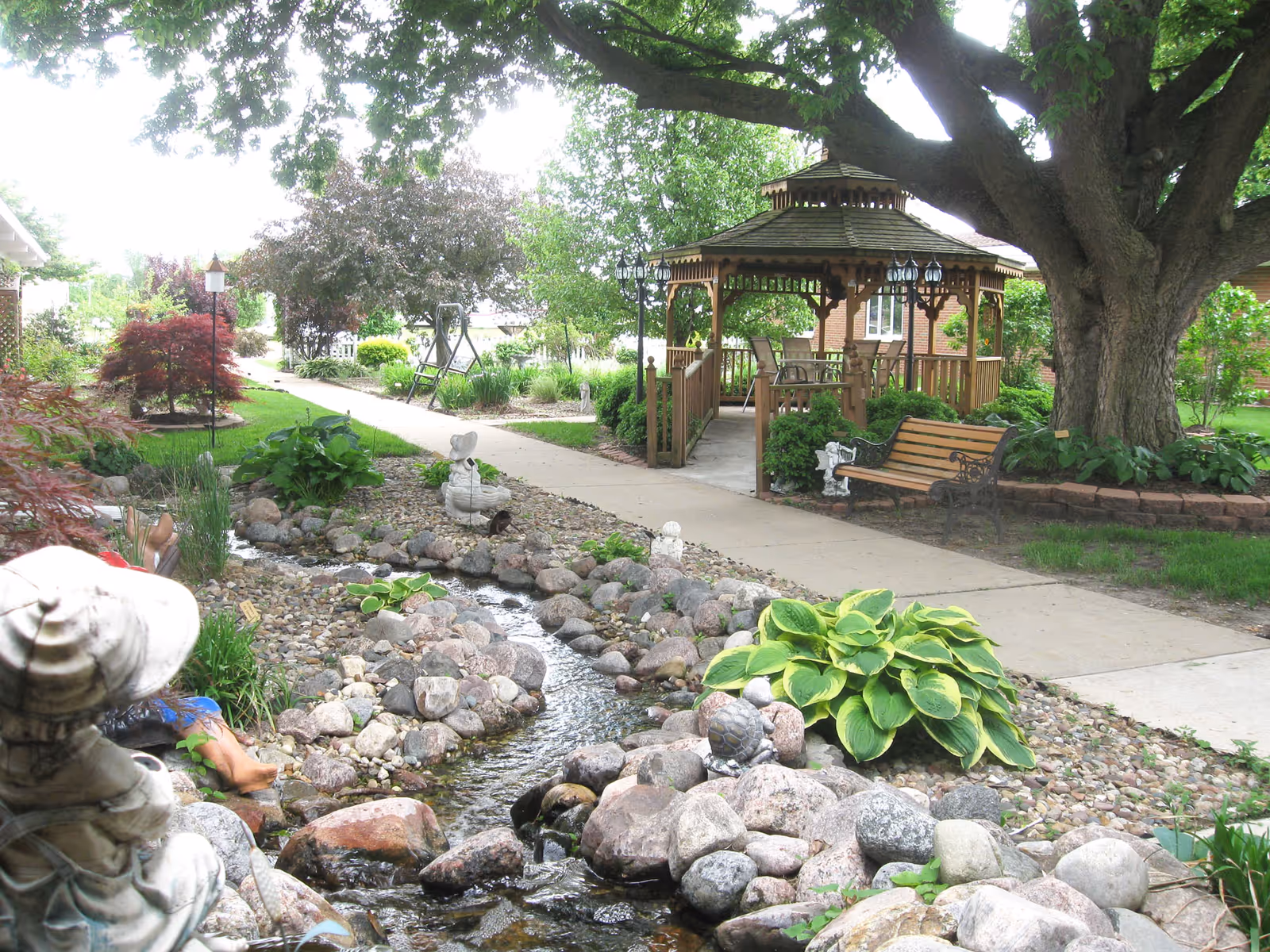 A peaceful outdoor garden area at a senior living facility featuring a small stream with rocks and plants along its banks. There is a paved walkway leading to a wooden gazebo with chairs inside. A large tree provides shade, and there is a wooden bench nearby. Various shrubs and decorative garden statues are also visible.