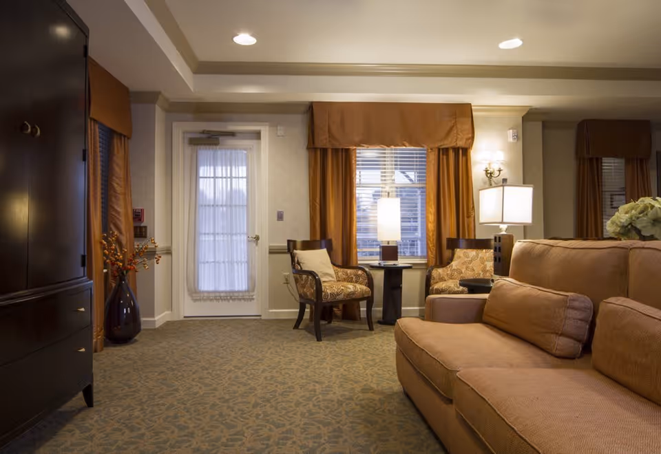 A cozy living room area with a large brown sofa and matching cushions, two patterned armchairs with a small round table and a lamp between them, a window with brown curtains, a door with a sheer curtain, a dark wooden cabinet on the left, and a vase with decorative branches on the floor.