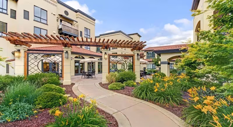 Outdoor garden area at Merrill Gardens at Campbell featuring a curved concrete pathway, lush green bushes, yellow flowers, and a pergola structure with seating tables and umbrellas near the building entrance.