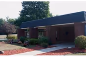 Single-story brick building with a covered entrance, concrete walkway, and landscaped shrubs and mulch beds.