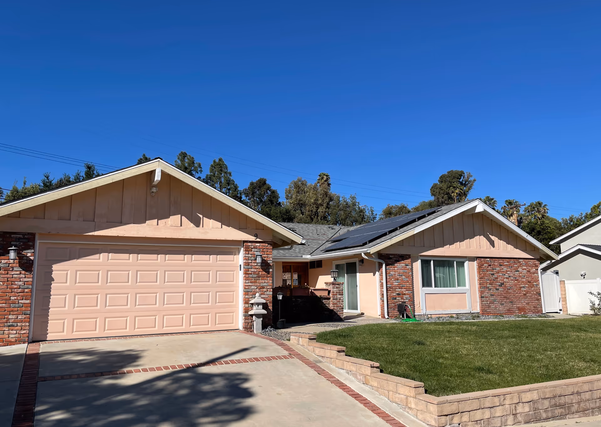 Single-story residential building with a two-car garage, brick and beige siding exterior, solar panels on the roof, a green lawn, and a clear blue sky.