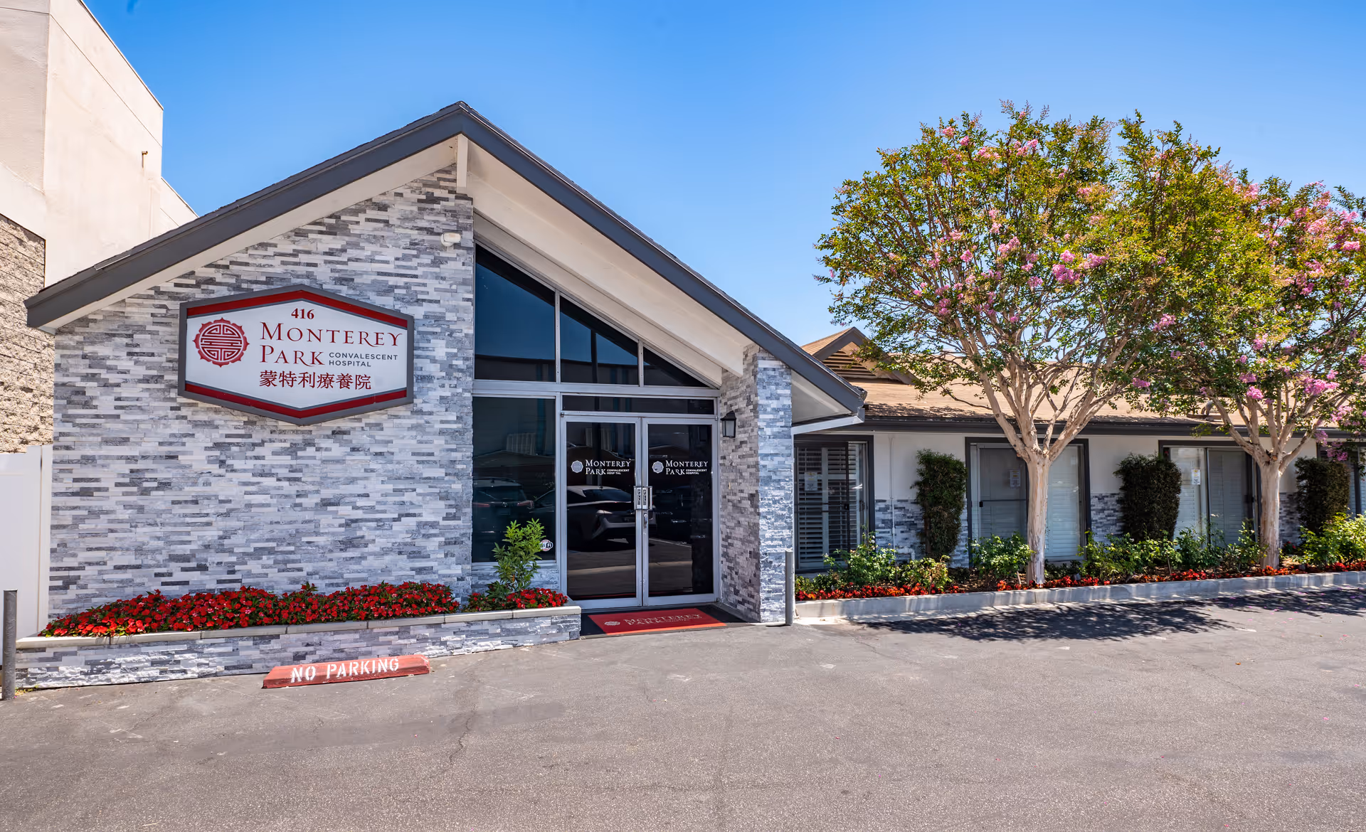 Front entrance of Monterey Park Convalescent Hospital with a stone-clad facade, glass double doors, red signage, and landscaped planters beneath flowering trees.