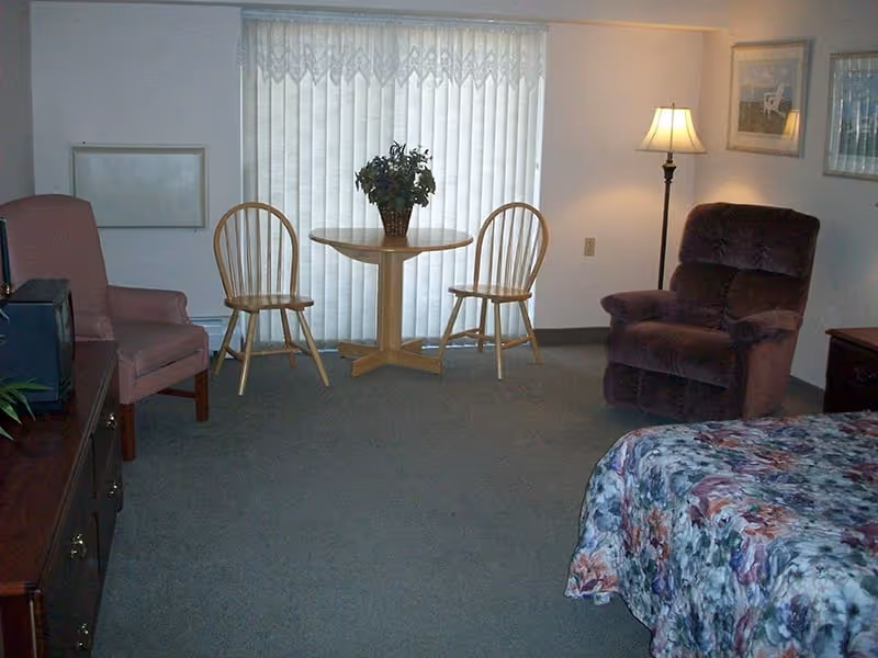 A cozy assisted living room with a floral bedspread on a bed in the foreground, a brown recliner chair, a wooden table with two wooden chairs, a floor lamp, and a pink armchair. The room has carpeted flooring and vertical blinds covering a window in the background.