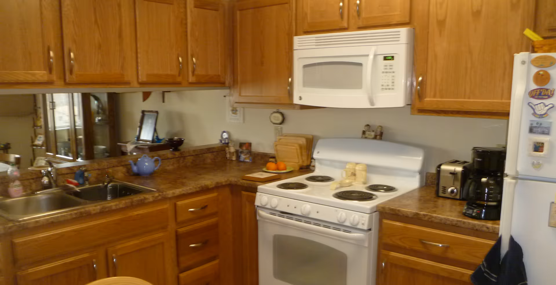 A kitchen with wooden cabinets, a white electric stove with four burners, a white microwave mounted above the stove, a double sink with a faucet, a toaster, a coffee maker, and a white refrigerator with magnets and decorations on the door. The countertops are brown with a marble pattern.