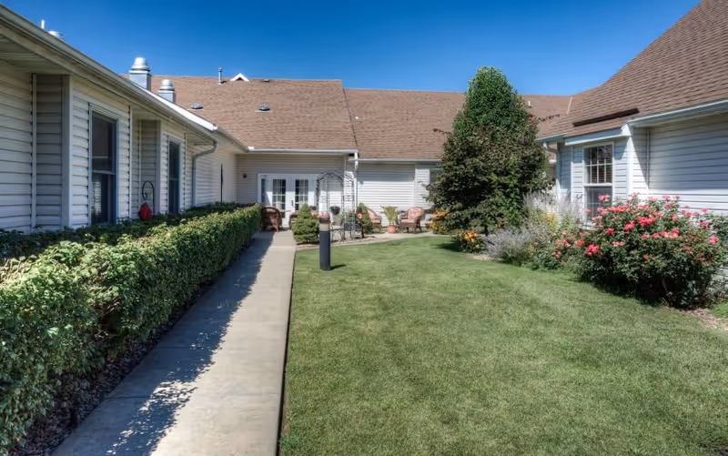Outdoor courtyard area at Vintage Park at Gardner with a concrete walkway, green grass, bushes, flowering plants, and patio furniture under a clear blue sky.