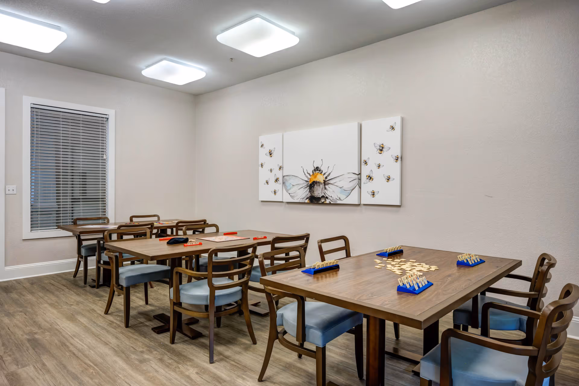 A well-lit room with three wooden tables and several chairs with blue cushions arranged around them. The tables have game tiles and racks set up, suggesting a game or activity area. On the wall, there is a three-panel artwork featuring a large bee and several smaller bees. The room has light-colored walls, wood flooring, and a window with closed blinds.