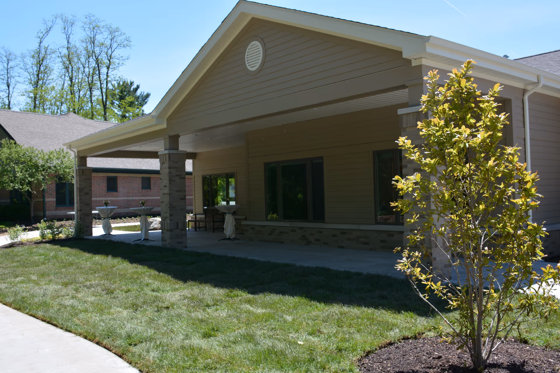 Exterior view of a covered patio area at a senior living facility with beige siding and brick pillars, surrounded by green grass and small trees under a clear blue sky.