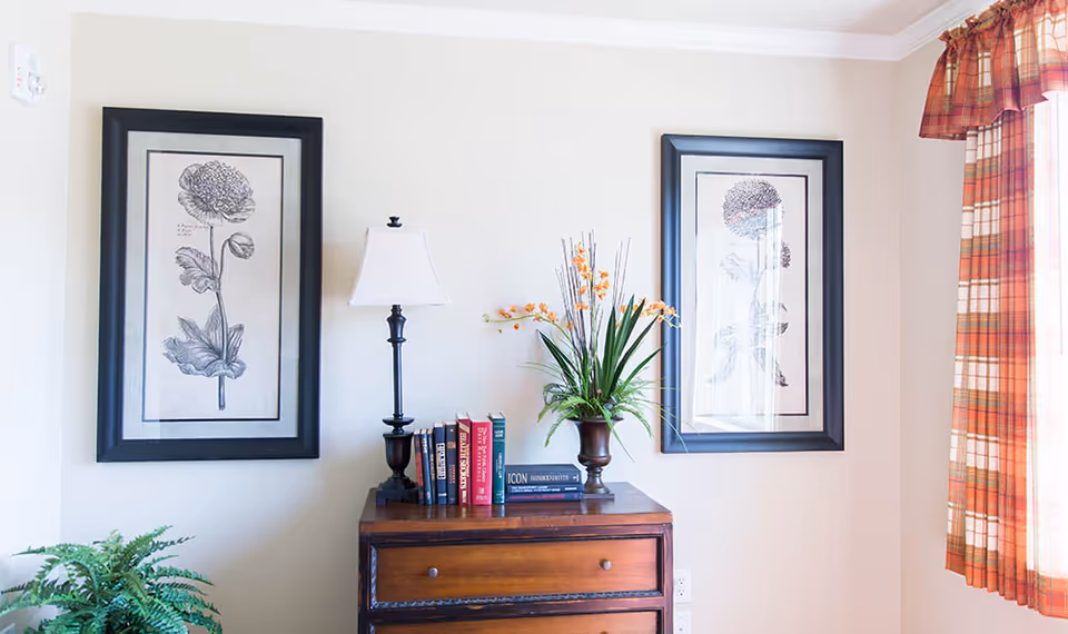 A corner of a room with a wooden chest of drawers topped with a table lamp, a small collection of books, and a vase with orange flowers. Two framed botanical prints hang on the wall above the chest. A window with red and orange plaid curtains is on the right side, and a green plant is partially visible on the left.